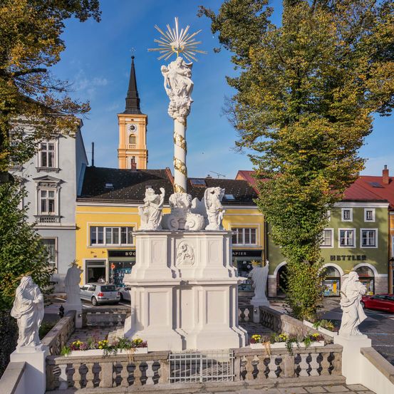 Eine weiße Statue steht in der Mitte eines Stadtplatzes, umgeben von Statuen und einem Geländer. Gebäude mit gelben und grünen Fassaden stehen dahinter. Bäume und ein Turm sind im Hintergrund zu sehen.