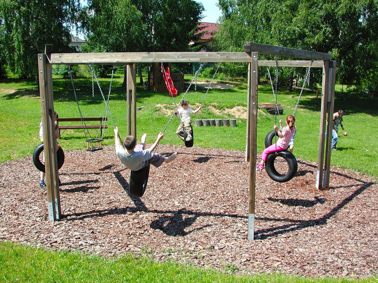 Three children play on swings in a grassy outdoor area. One swings from a tire, another from a chain, and a third from a slide.