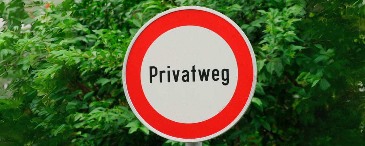 Ein weißer runder Schild mit rotem Rand zeigt das Wort 'Privatweg'. Hinter dem Schild befinden sich grüne buschige Büsche.