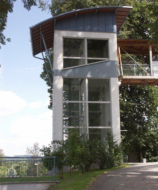 Ein modernes, eckiges Gebäude mit einer Glaswand, einer Holzterrasse und einem Metallgeländer. Das Gebäude ist von Bäumen und Pflanzen umgeben.