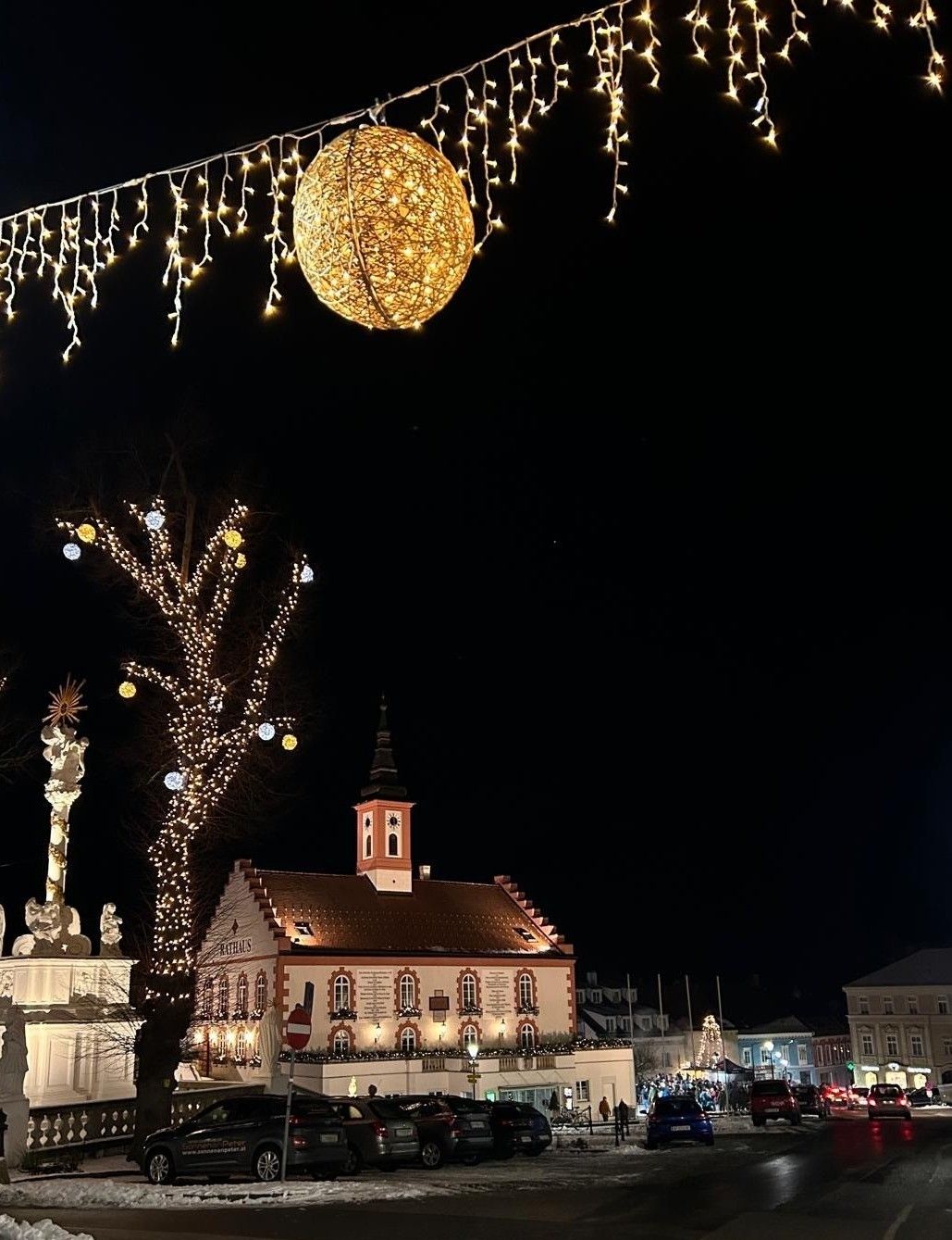 Eine nächtliche Szene eines Stadtplatzes mit festlichen Dekorationen. Eine große beleuchtete Kugel hängt darüber, während ein Uhrturm neben einem wunderschön dekorierten Gebäude steht.