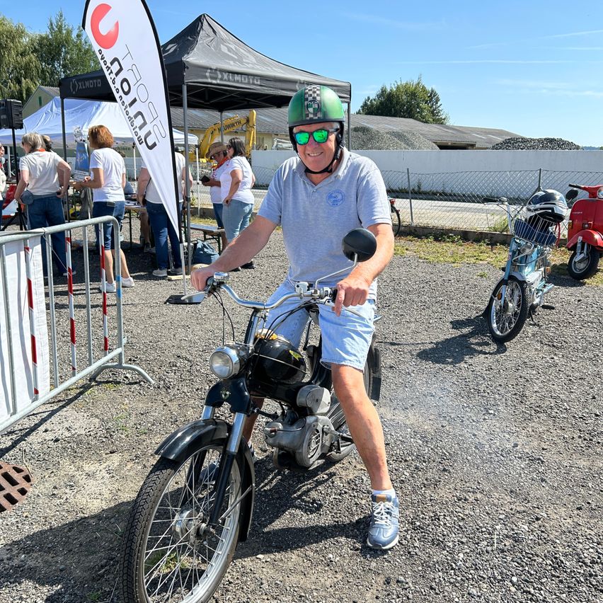 Ein Mann mit Helm und Sonnenbrille sitzt auf einem Motorrad im Freien. Hinter ihm stehen mehrere Personen in der Nähe eines Zeltes und eines weißen Zauns. In der Ferne sind weitere Personen und ein Baufahrzeug zu sehen.