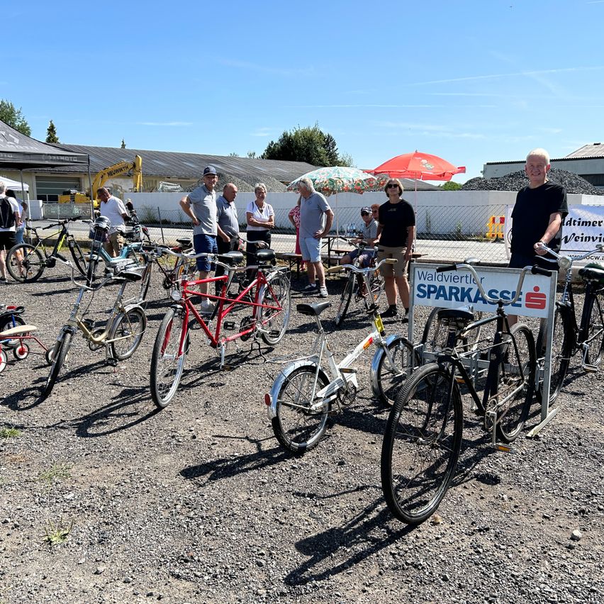 Eine Gruppe von Menschen steht um verschiedene Fahrräder auf einem Schotterplatz, wobei ein Mann in einem schwarzen T-Shirt ein Fahrrad mit einem Schild 'Waldviertler Sparkasse' fährt.