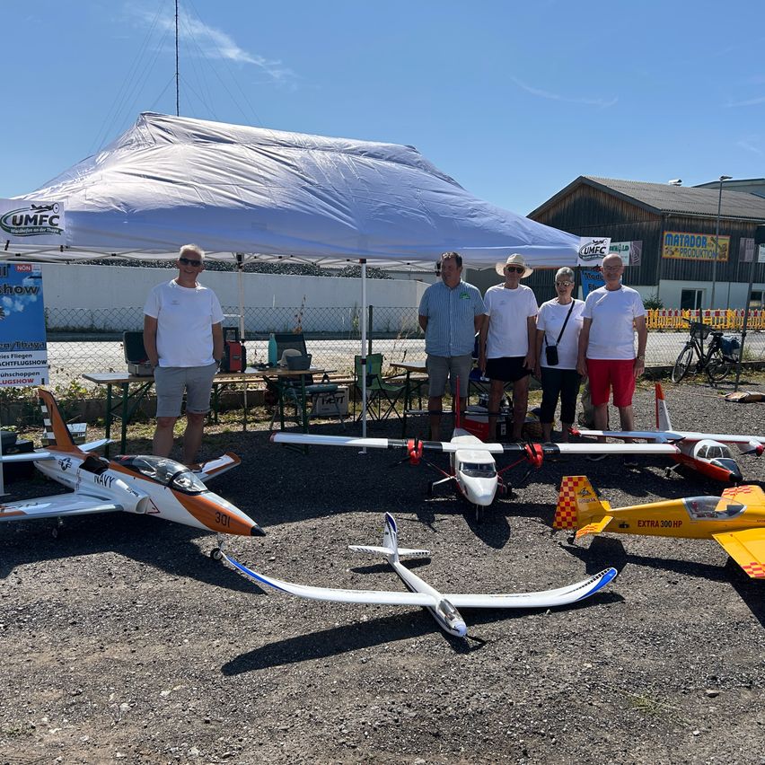 Fünf Personen stehen unter einem Zelt mit mehreren ausgestellten Modellflugzeugen vor ihnen. Die Modelle sind in verschiedenen Farben und Größen.