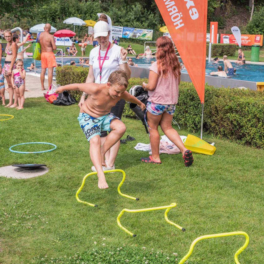 Ein junger Junge springt über gelbe Hürden auf einem Rasenfeld, während eine Frau einen Beutel hält. In der Nähe gehen eine Frau mit Hut und ein Mädchen. Im Hintergrund befindet sich ein Pool mit schwimmenden Menschen und ein Banner mit dem Text 'KÖNIG'.