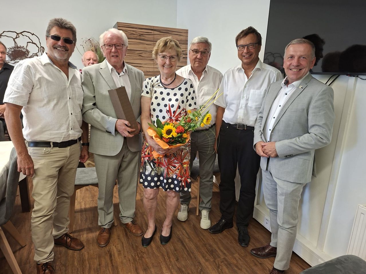 A group of six people are standing together in a room, smiling and posing for a photo. The woman in the center is holding a bouquet of flowers.