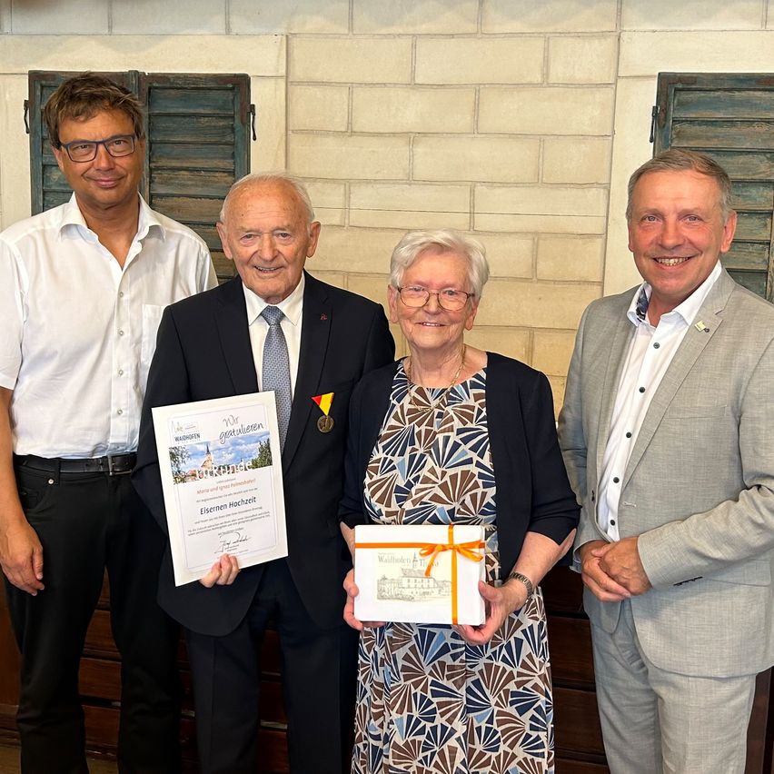 Four individuals stand together, smiling, with two of them holding certificates and gifts. The man on the left wears glasses and a belt. The woman in the center wears glasses and a necklace. The man on the right wears a suit and a medal.