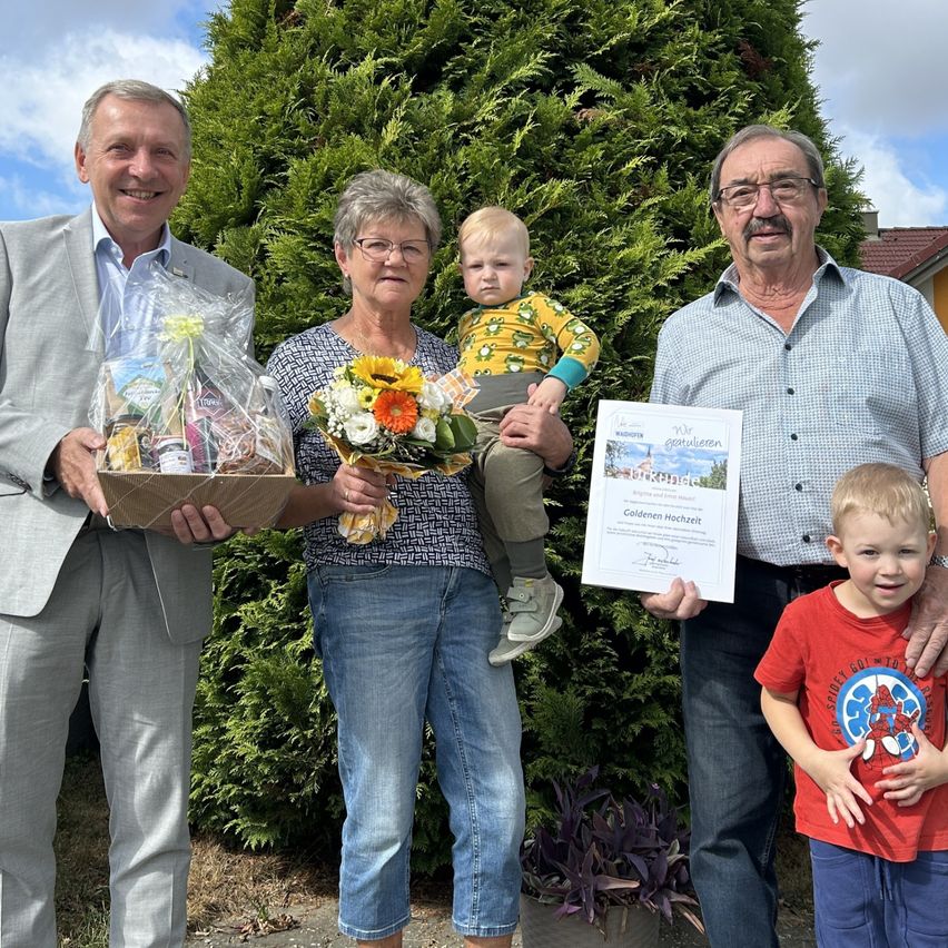 A family of four adults and two children stands in front of a hedge, smiling. The man on the left holds a gift basket, the woman in the middle holds flowers, and the man on the right holds a certificate.