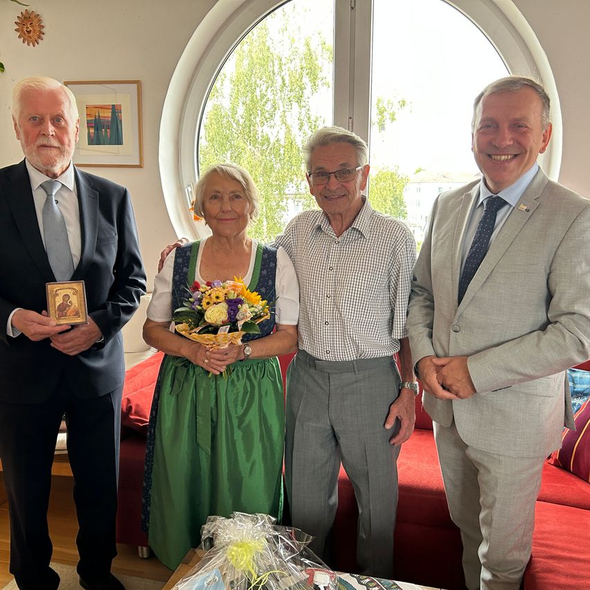Four individuals are standing together in a room, smiling for a photo. A woman holds a bouquet of flowers, and a man on the left holds a small icon. The man on the right wears a pin on his jacket.