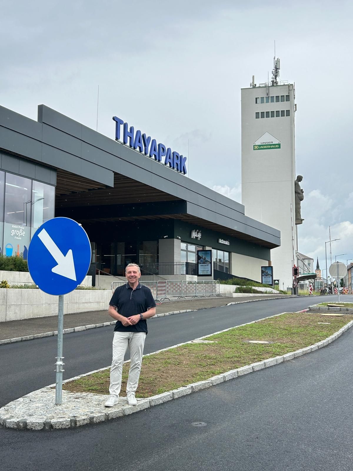 Ein Mann in Freizeitkleidung steht vor dem Thaypark-Gebäude, einem großen Bauwerk mit einem hohen Turm im Hintergrund. Ein blaues Straßenschild mit einem Pfeil zeigt nach rechts.