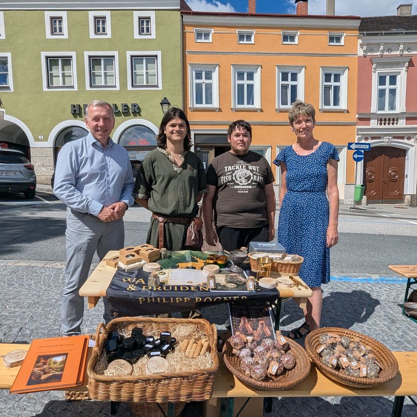 Vier Personen stehen hinter einem Tisch mit verschiedenen Gegenständen, darunter Körbe mit Kräutern, einem Buch und Gläsern. Der Mann links trägt ein hellblaues Hemd. Hinter ihnen befinden sich Gebäude, darunter eines mit dem Wort 'HELLER' darauf.