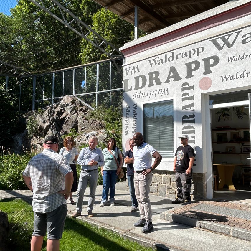 Eine Gruppe von Menschen steht vor einem Gebäude namens Waldrapp. Sie unterhalten sich und einige schauen auf das Gebäude.