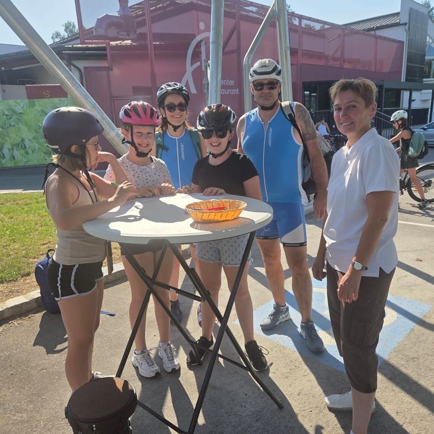 A group of people, some wearing helmets, stand around a table with a basket on it. They are smiling and appear to be posing for a photo. Behind them, there is a building with a red wall and a person riding a bicycle.