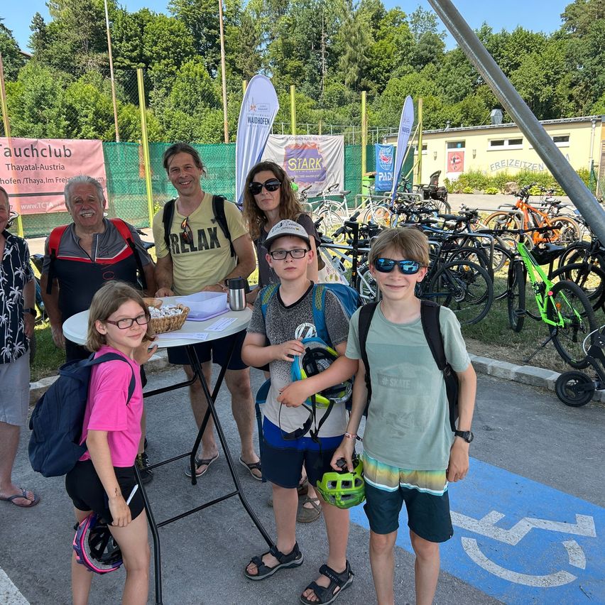 A group of people, including adults and children, stand in front of a table with a basket of cookies and bicycles parked behind them. They are all smiling and appear to be posing for a photo.