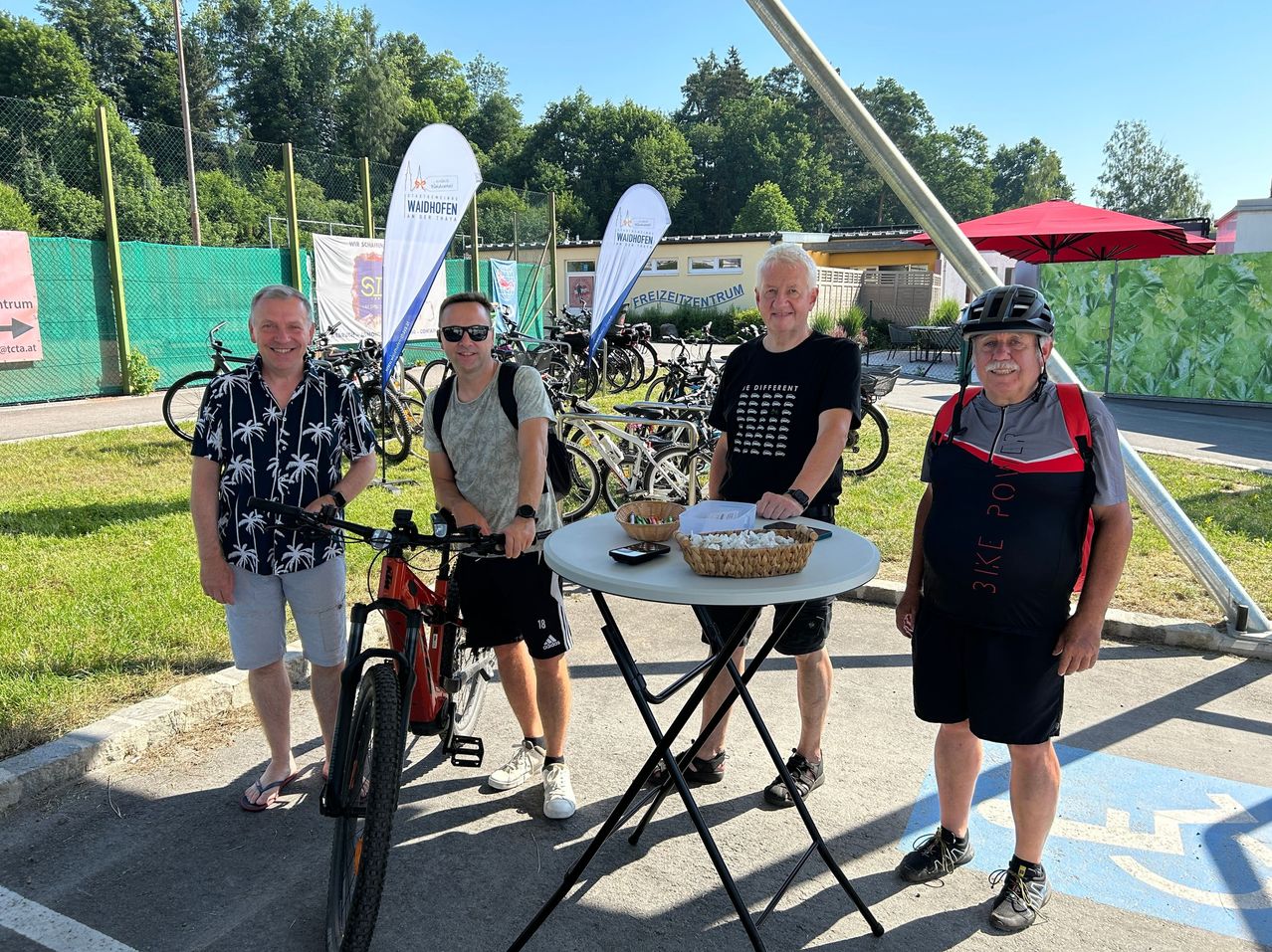 Four men stand next to a table with a basket of snacks, smiling and posing for a photo. Behind them, a row of bicycles is parked, and banners are displayed. Trees and a building are in the background.