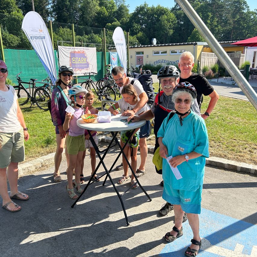 A group of people in cycling gear are gathered around a table with food, possibly at a cycling event. Bicycles are parked in the background.
