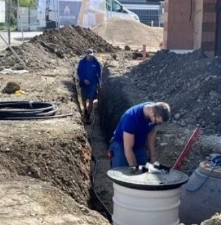 Zwei Arbeiter in blauen Overalls stehen in einer Baugrube, einer gebeugt und der andere stehend, mit einem Haufen Erde dahinter. Ein Gebäude, ein Van und ein Eimer sind im Hintergrund zu sehen.