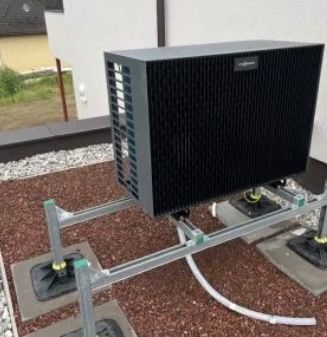 Eine Außenklimaanlage ist an einem Metallrahmen montiert, mit angeschlossenen Rohren und Kabeln, vor einer weißen Wand.