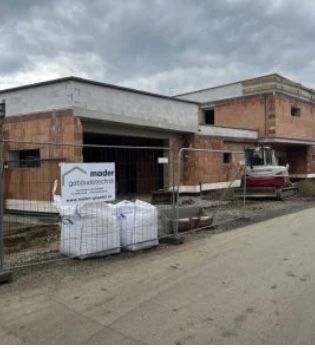 Ein Baugelände mit einem teilweise fertiggestellten Gebäude, einem Schild für die Baufirma 'moder' und geparkten Baumaschinen davor