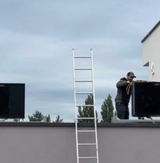 Ein Mann steht auf einem Dach und hält ein schwarzes Gerät, wahrscheinlich eine Klimaanlage. Er trägt einen Hut und eine Jacke. Eine Leiter steht auf dem Dach. Im Hintergrund sind Bäume und ein bewölkter Himmel zu sehen.