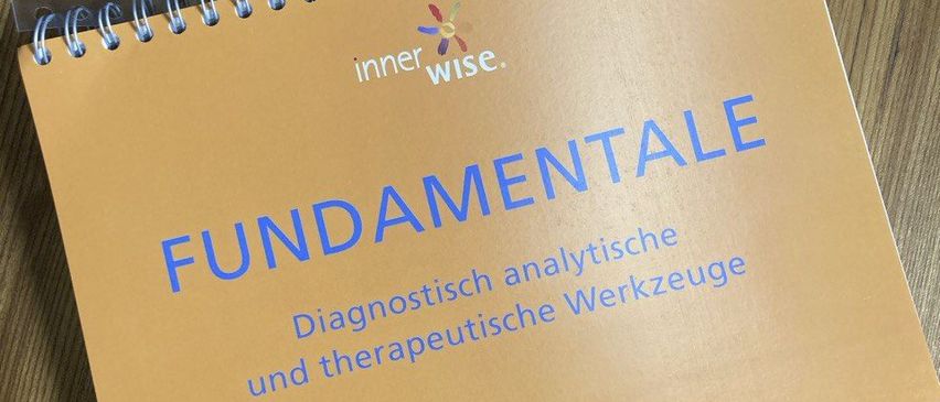 Ein braunes Spiralnotizbuch mit dem Titel 'FUNDAMENTALE' von Uwe Albrecht, verwendet für diagnostische und therapeutische Werkzeuge, liegt auf einem Holztisch.