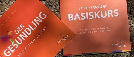 Zwei rote Bücher mit den Titeln "Gesundlich" und "Innerwise Basiskurs" liegen auf Kieselsteinen, darunter verstreut sind Karten mit farbigen Kreisen und dem Wort "Ja".