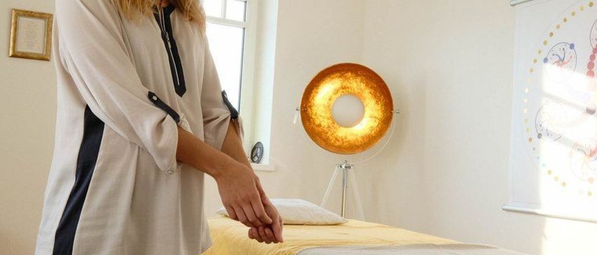 Eine Frau mit blonden Haaren steht in einem Zimmer und bereitet ein Bett mit einem gelben Laken und einer weißen Decke vor. Eine Lampe steht auf dem Boden und ein Poster hängt an der Wand.