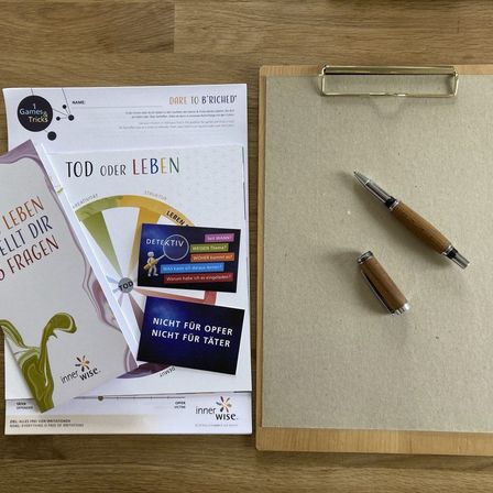 Ein Klemmbrett mit einem Stift, einem Prospekt mit dem Titel Tod oder Leben und einem weiteren Prospekt mit dem Titel 1 Spiele und Tricks auf einer Holzoberfläche.