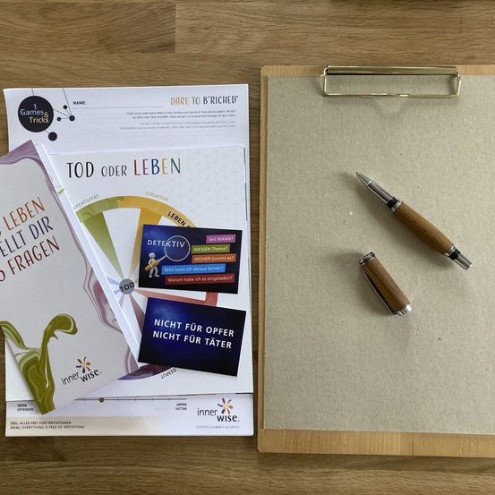 Ein Klemmbrett mit einem Stift, einem Prospekt mit dem Titel Tod oder Leben und einem weiteren Prospekt mit dem Titel 1 Spiele und Tricks auf einer Holzoberfläche.