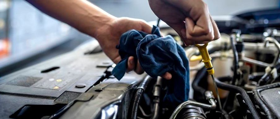 Ein Mechaniker arbeitet in einer Garage an einem Automotor, hält einen Schraubendreher und ein blaues Tuch in der Hand, mit einem Fenster und Gegenständen im Hintergrund.