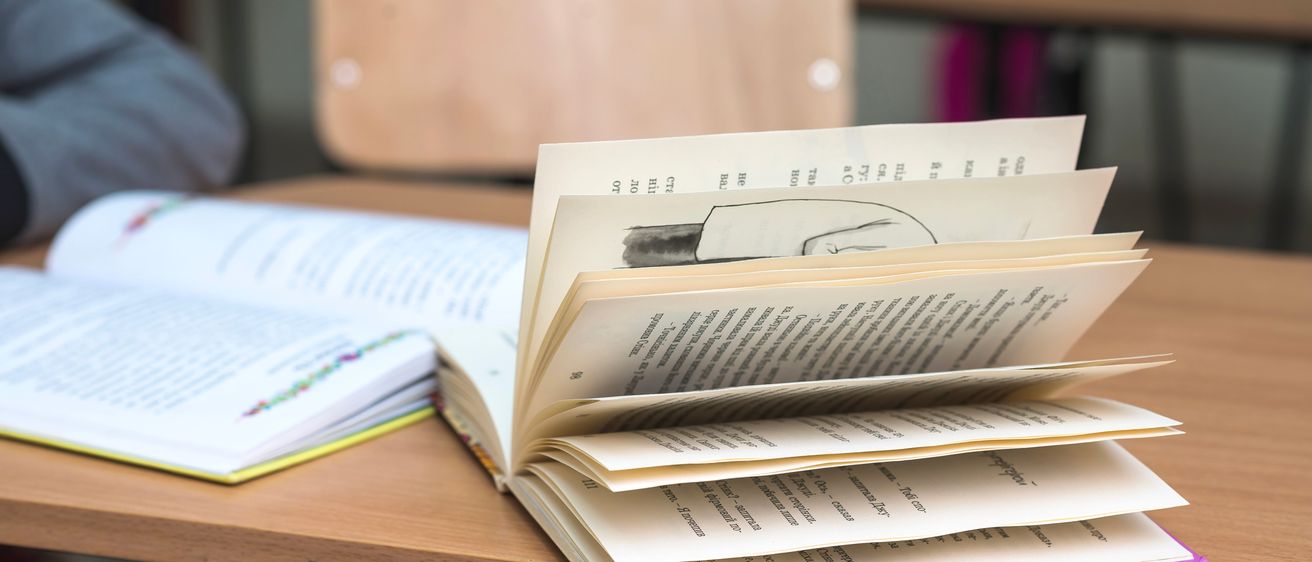 Ein offenes Buch mit einer Zeichnung auf seiner Seite liegt auf einem Holztisch in einem Klassenzimmer. Ein Stuhl ist im Hintergrund zu sehen.