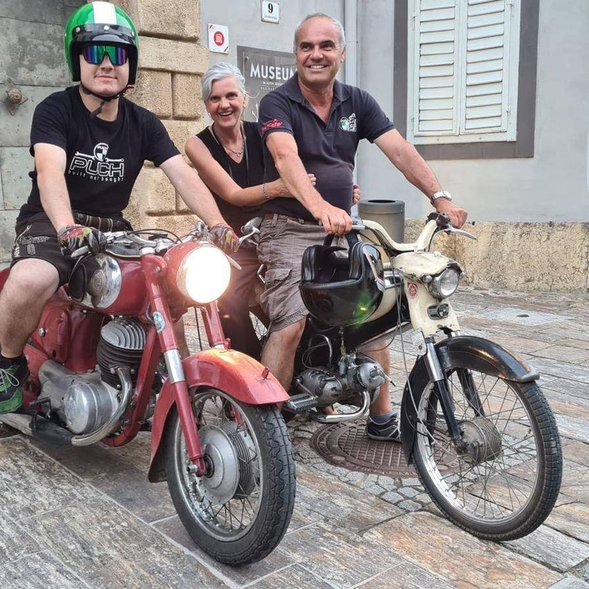 Drei Personen posieren mit ihren Motorrädern vor einem Gebäude mit der Aufschrift 'Museum.' Der Mann links trägt einen Helm und ein schwarzes T-Shirt. Die Frau in der Mitte trägt eine Halskette. Der Mann rechts trägt eine Uhr und ein blaues Poloshirt.