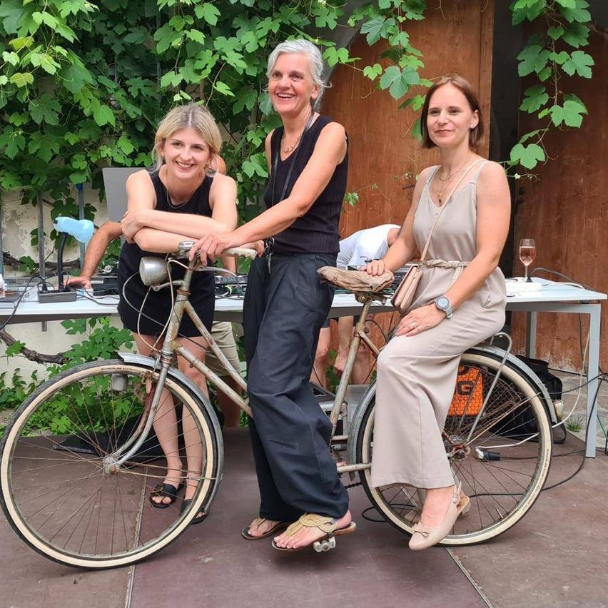 Drei Frauen posieren auf einem Fahrrad im Garten. Eine in der Mitte mit einer Halskette und hält das Lenkrad. Hinter ihnen steht ein Tisch mit einem Weinglas und einer Lampe.