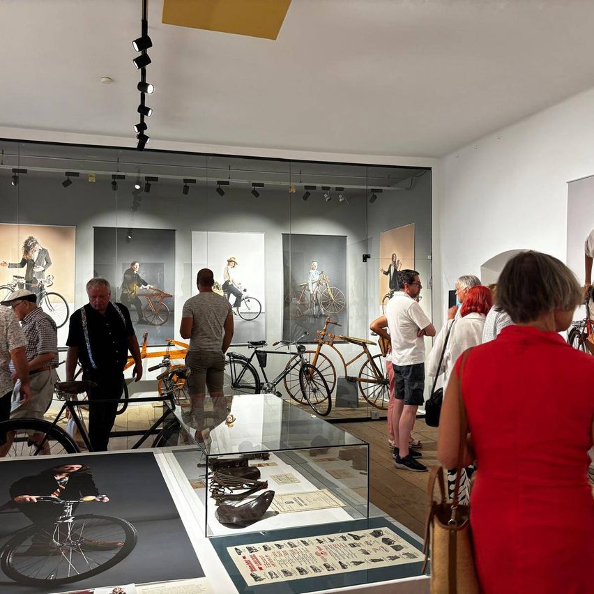 Eine Gruppe von Menschen betrachtet Fahrräder in einer Galerie. Die Fahrräder sind an den Wänden ausgestellt, und ein Glaskasten zeigt Fahrradteile. Eine Frau in einem roten Kleid steht vorne.