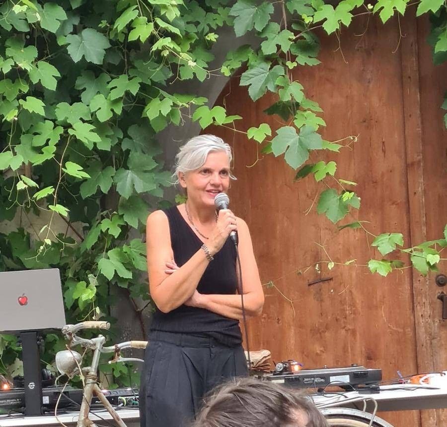 Eine Frau steht mit einem Mikrofon in der Hand vor einer mit grünem Laub bedeckten Holzwand. Sie trägt ein schwarzes ärmelloses Oberteil und eine Halskette. Hinter ihr sind ein Fahrrad und elektronische Geräte zu sehen.