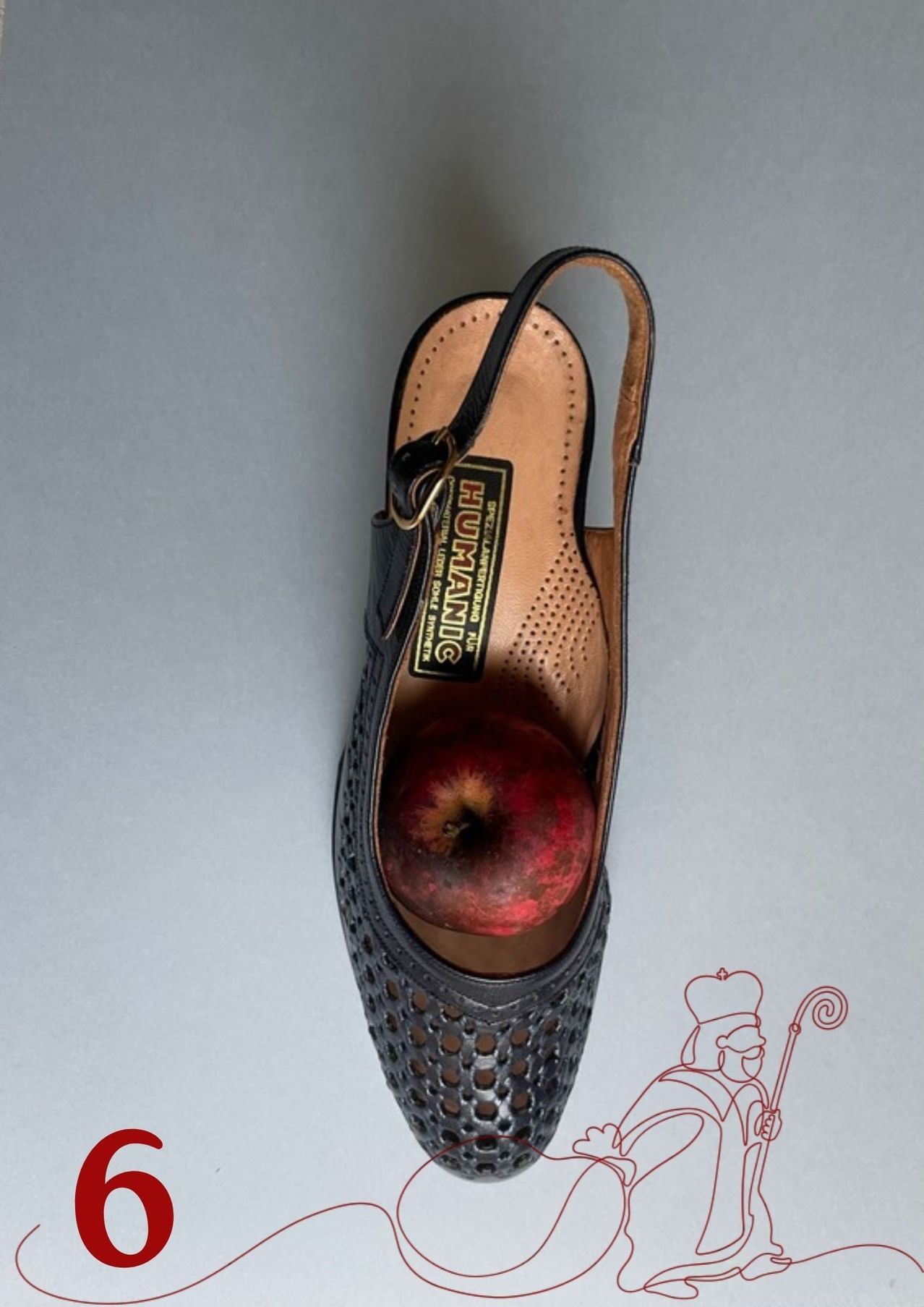 Ein schwarzer Sandalenschuh mit Riemen liegt auf einer grauen Oberfläche. Ein Apfel befindet sich im Inneren des Schuhs. Der Schuh hat ein Markenlabel mit der Aufschrift HUMANIC. Auf der rechten Seite ist eine Karikatur eines Mannes mit Krone und Stab zu sehen.