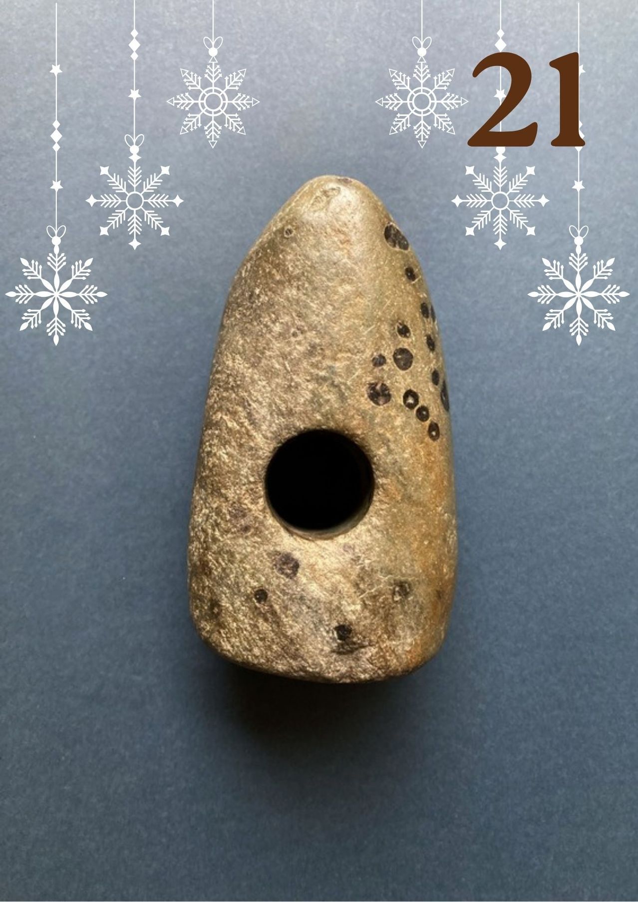 Ein verwittertes Steinobjekt mit einem Loch sitzt auf einem grauen Hintergrund, umgeben von weißen Schneeflockendekorationen.