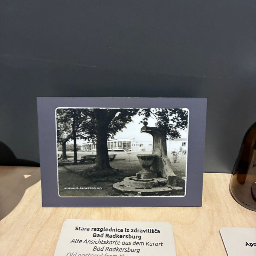A framed old postcard of Bad Radkersburg, an old health resort town, displayed on a wooden table. The image features a fountain, trees, and benches.