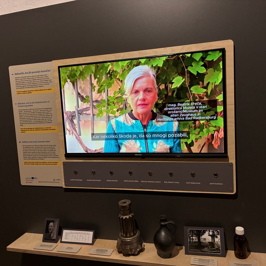 A monitor displays a video with a woman, text in Croatian. Below, there are several buttons, a photo frame, a bottle, and a jug on a wooden shelf.