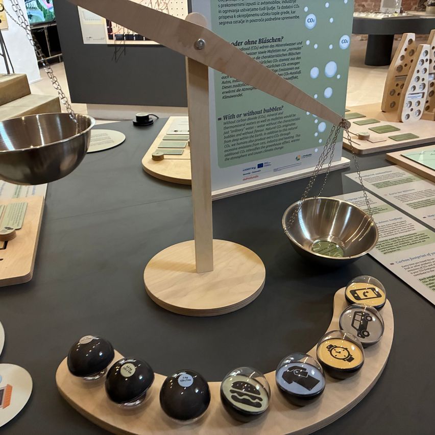 A balance scale displays two metal bowls. A wooden arm holds up an informational sign about CO2 emissions. Below are various objects representing carbon footprints.