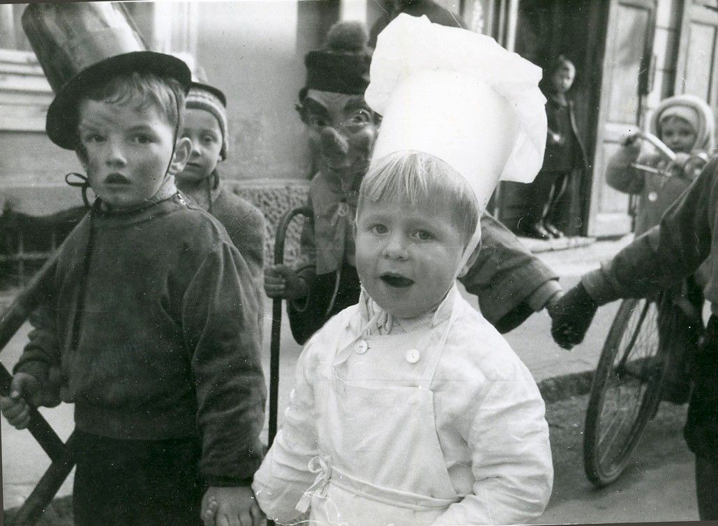 Ein Schwarz-Weiß-Foto eines Kindes in einer Kochmütze, das neben einem anderen Kind und einer vermaskierten Person steht. Hinter ihnen hält eine Person einen Stock.