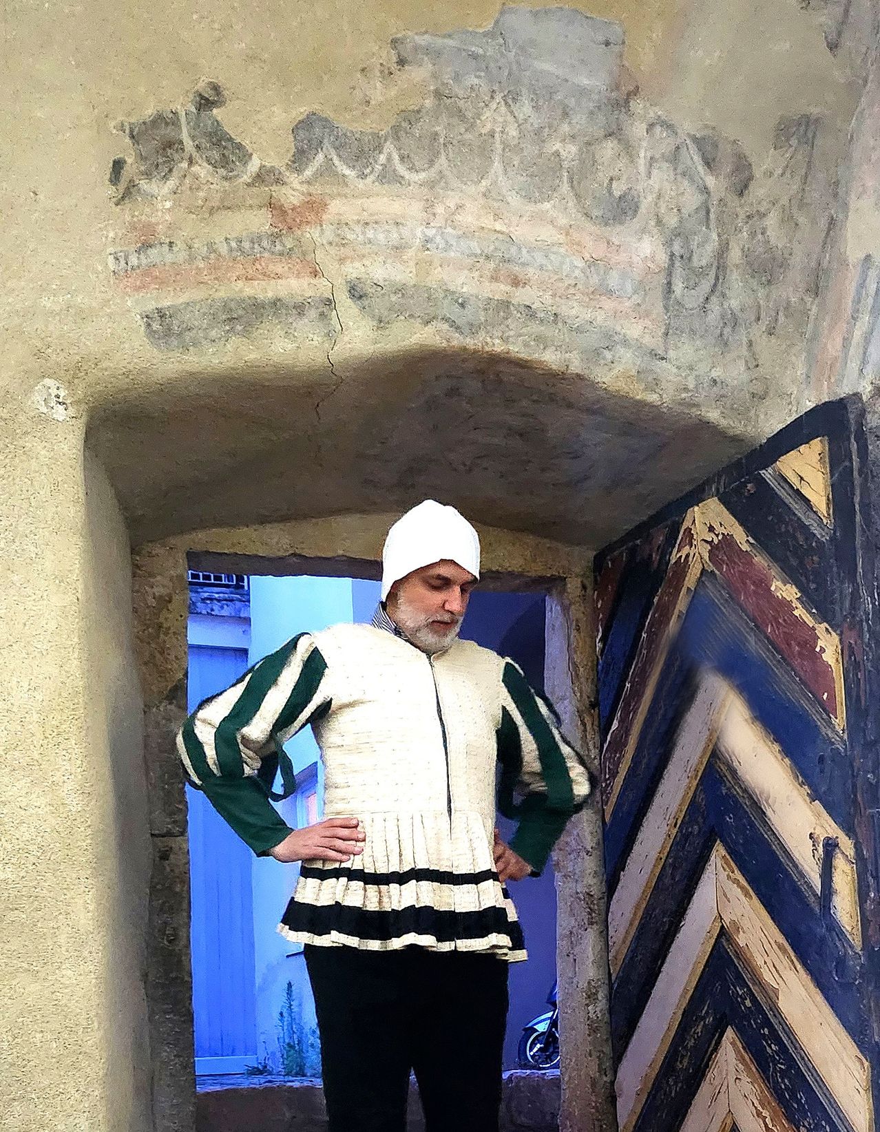 Ein Mann in einem weißen und grünen historischen Kostüm steht in einem Bogengang mit einer verwitterten Steinmauer und einer bunten Holztür.