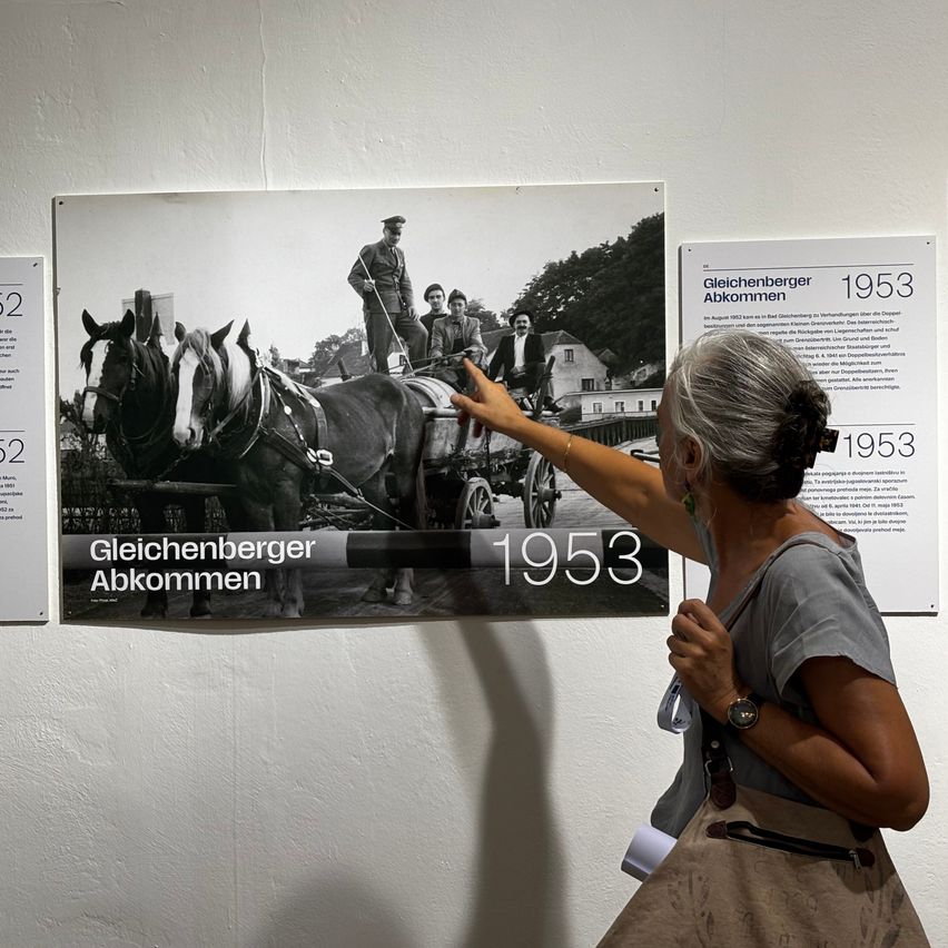 Eine Frau zeigt auf ein Schwarz-Weiß-Foto einer Pferdekutsche aus dem Jahr 1953, das an einer Wand neben anderen Informationsplakaten hängt.