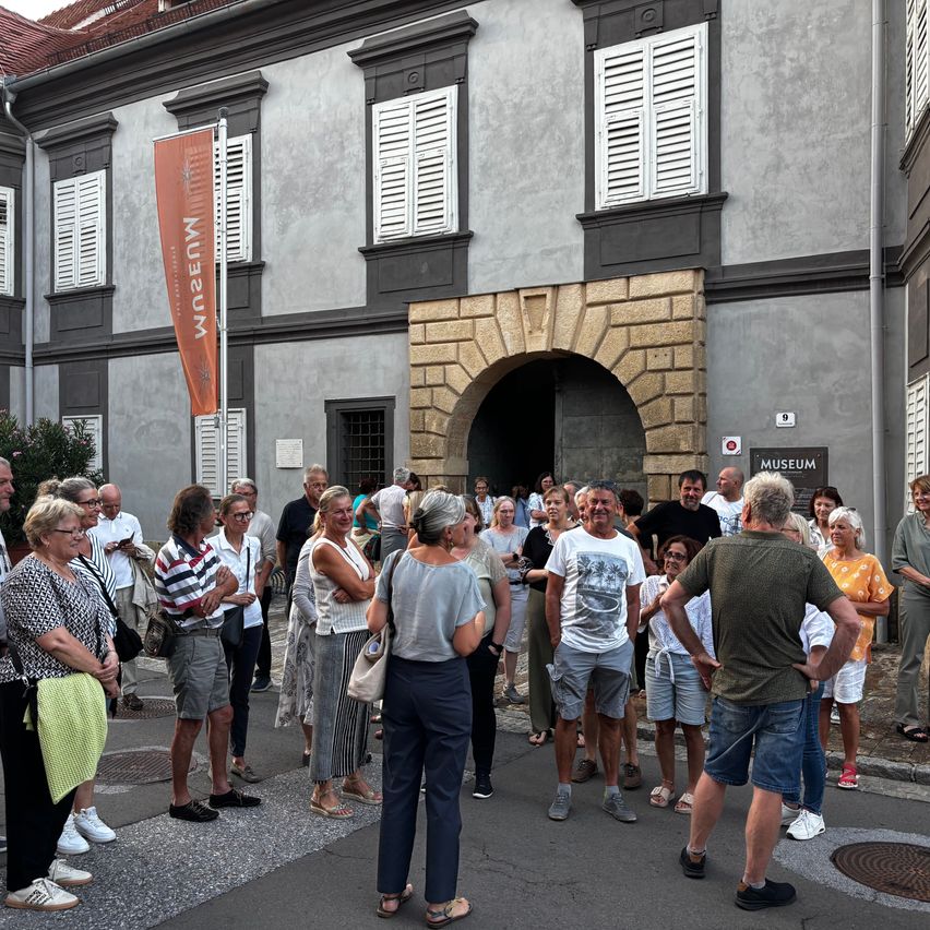 Eine Gruppe von Menschen, wahrscheinlich Touristen, versammelt sich vor einem Gebäude mit einem gewölbten Eingang und einer Fahne. Das Gebäude hat mehrere Fenster und ein Schild über dem Eingang.