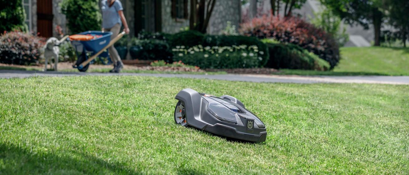 Eine Frau schiebt eine blaue Schubkarre in einem Hinterhof mit einem Roboter-Rasenmäher im Vordergrund.