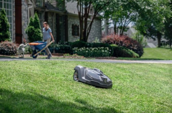 Eine Frau schiebt eine Schubkarre, während ein robotergesteuerter Rasenmäher auf dem Rasen in der Nähe eines Hauses ruht.
