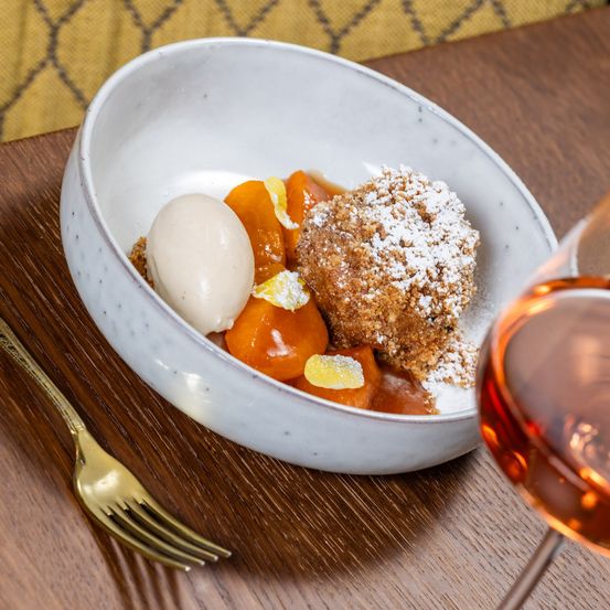 Eine weiße Schüssel mit einem Dessert darin, bestehend aus Pfirsichscheiben, einer Vanilleschote und einem knusprigen Keks. Daneben steht ein Weinglas und eine goldene Gabel auf einem Holztisch.