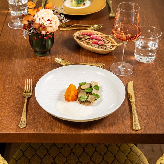 Ein wunderschön gedeckter Esstisch mit einem weißen Teller mit Essen, einem Weinglas, Besteck und einer dekorativen Blumenarrangement auf einer Holzoberfläche.