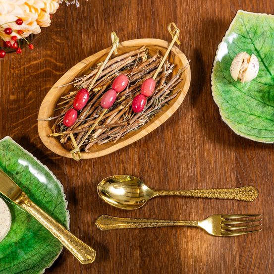 Auf einem Holztisch steht eine Schale mit Spießen aus rotem Essen, ein Teller mit Blattdesign und Essen, eine goldene Gabel und ein goldener Löffel.