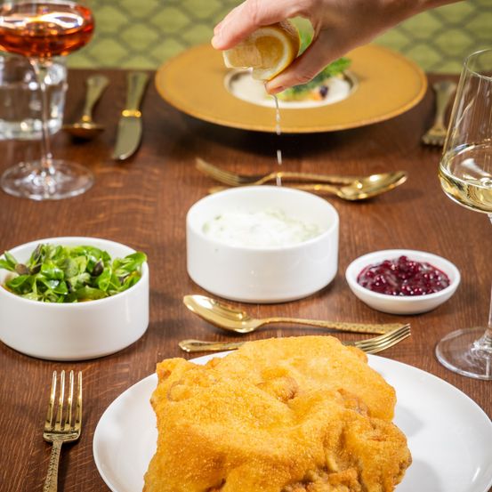 Ein Holztisch ist mit einem goldenen Teller, einer Schüssel Salat, einer Schüssel saure Sahne, einer Schüssel Cranberries, Weingläsern, Besteck und einem gebratenen Gericht auf einem weißen Teller gedeckt.